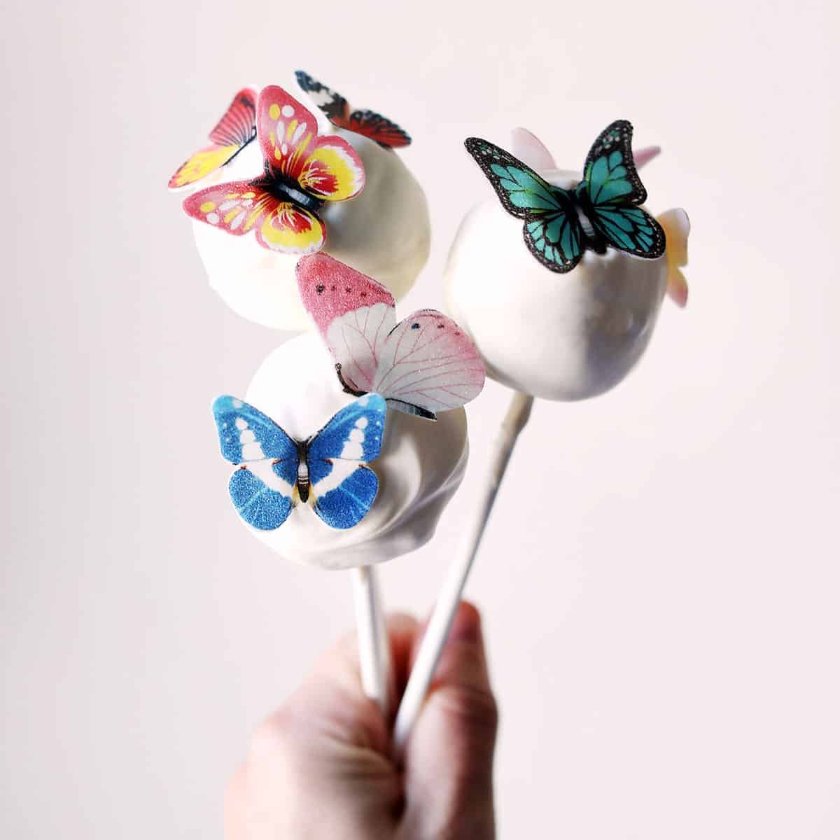 someone holding 3 white cake pops with fake butterflies on top