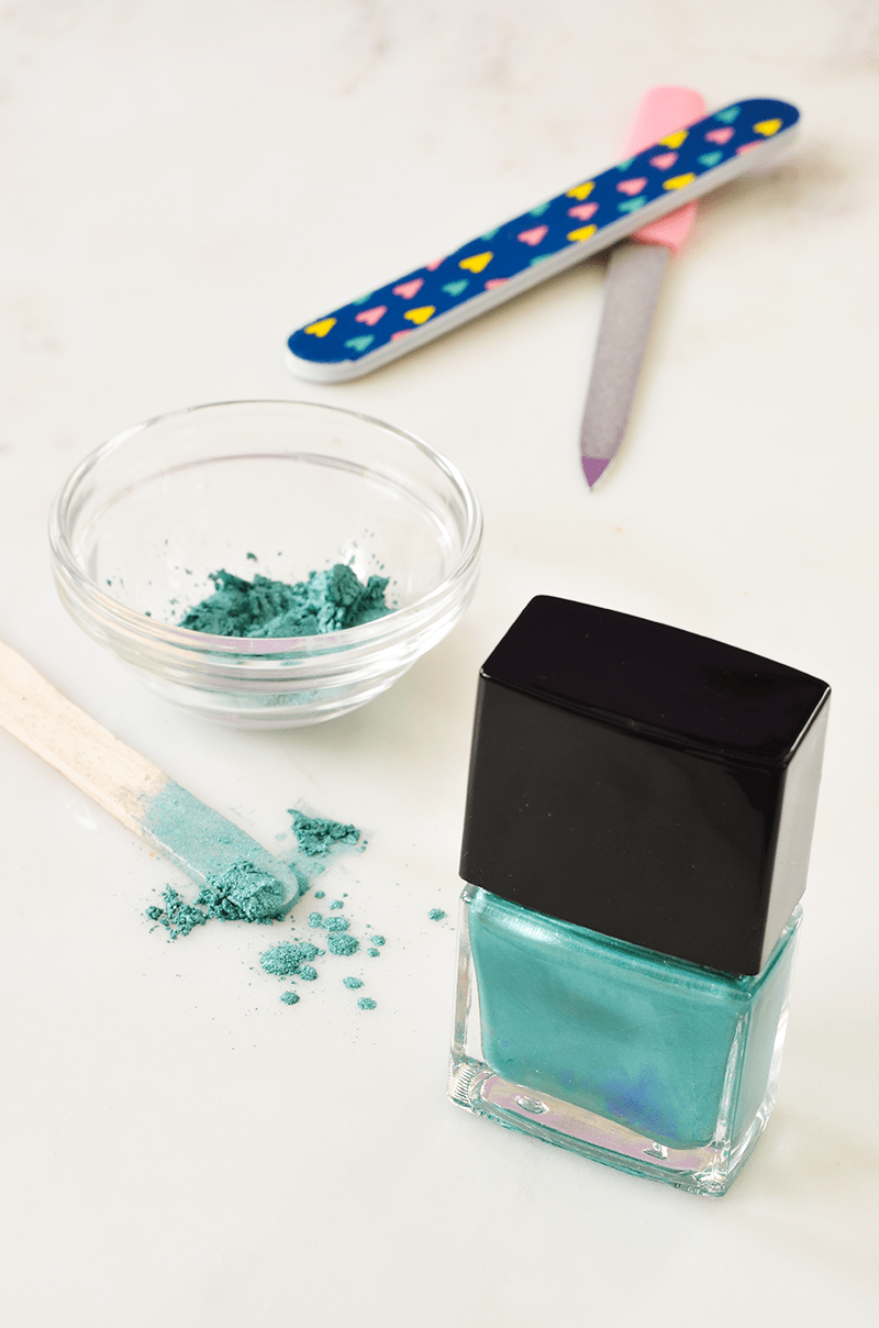 teal nailpolish in a jar with teal powder in a clear bowl and on a popcycle stick with 2 nail files by them