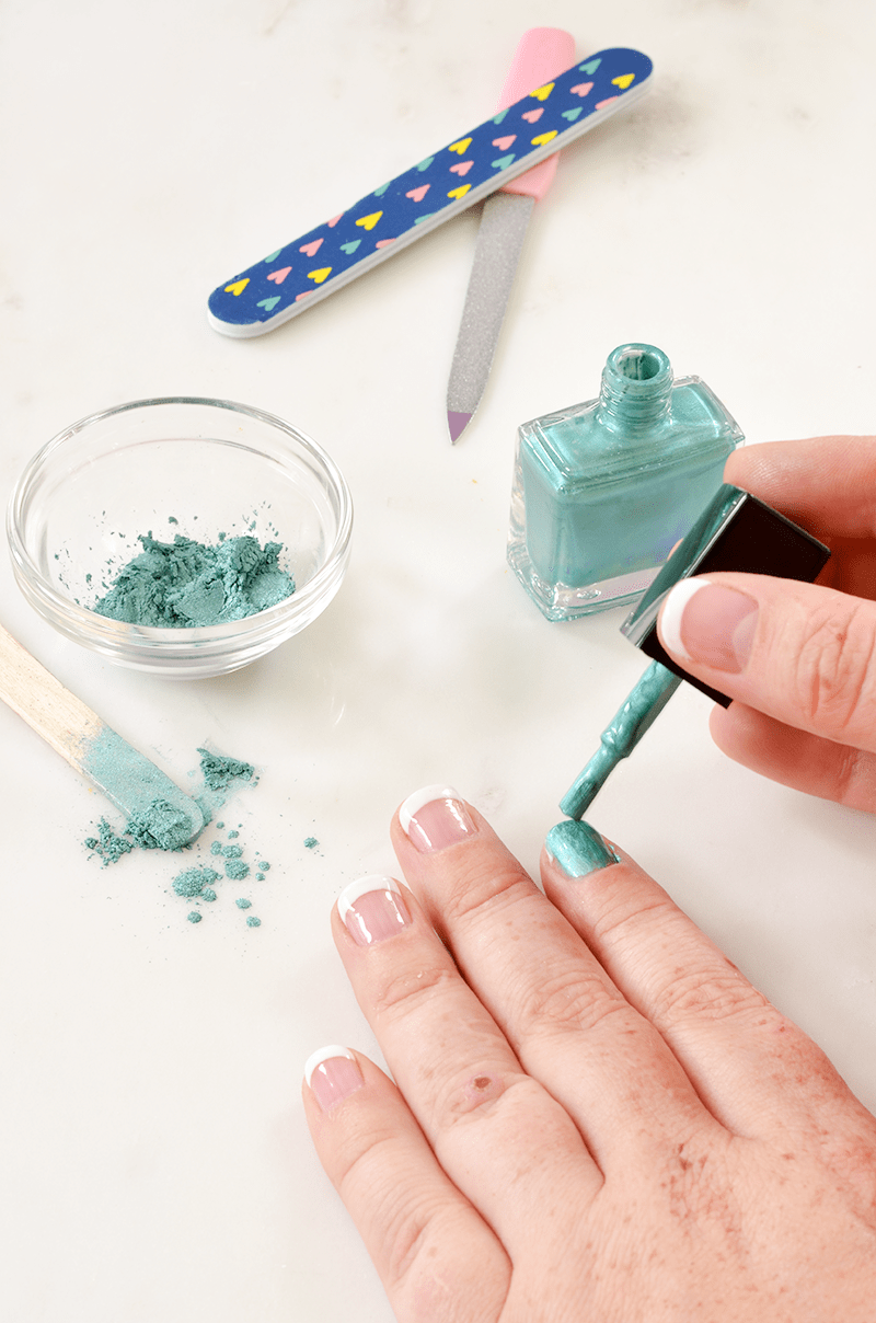 someone painting their pointer nail with teal nail polish with a jr of teal powder in a clear bowl, a popcycle stick, and 2 nail filers next to them
