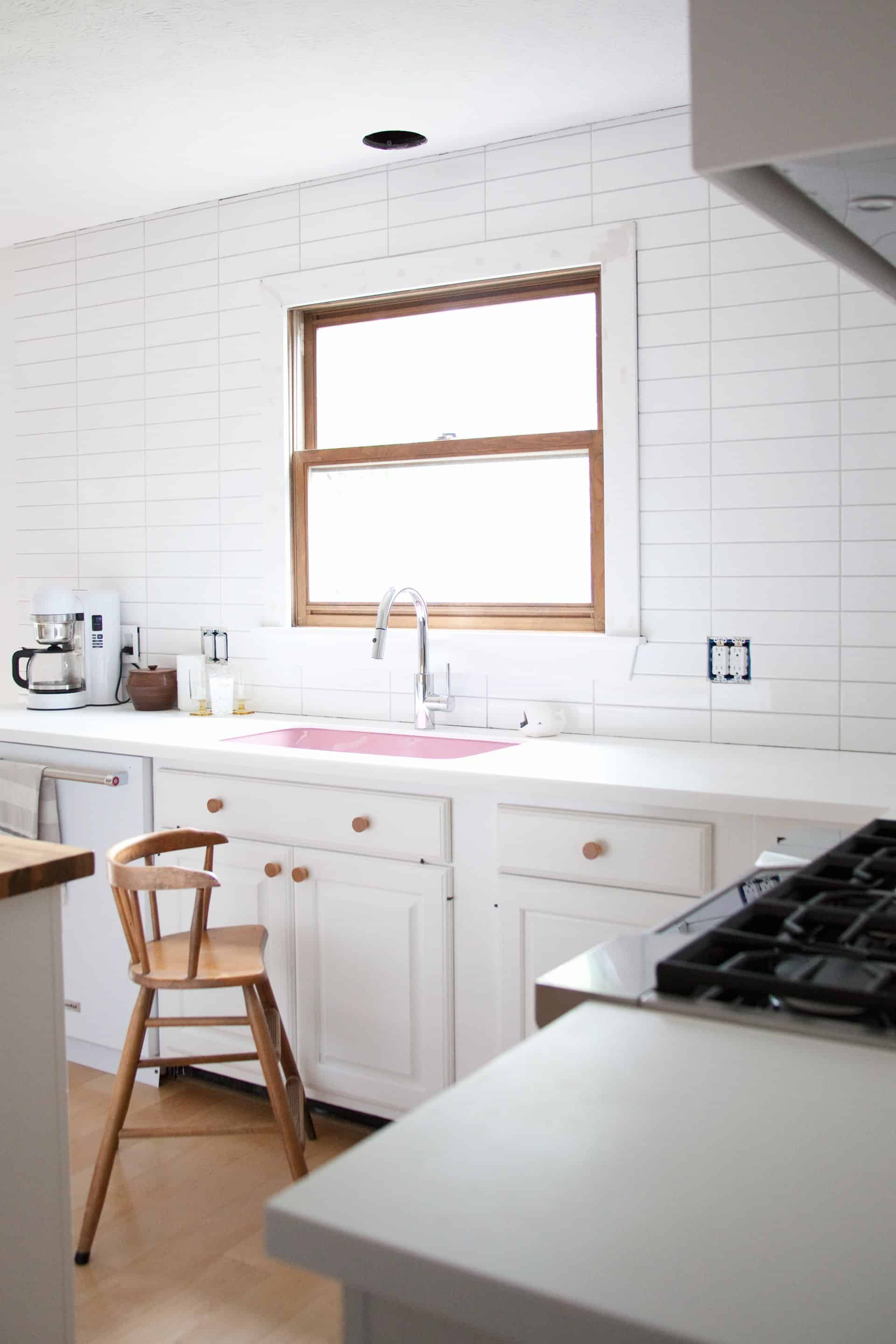 a kitchen with white cabinets with wooden handles, white countertops, and white tile with a stool in front of the sink
