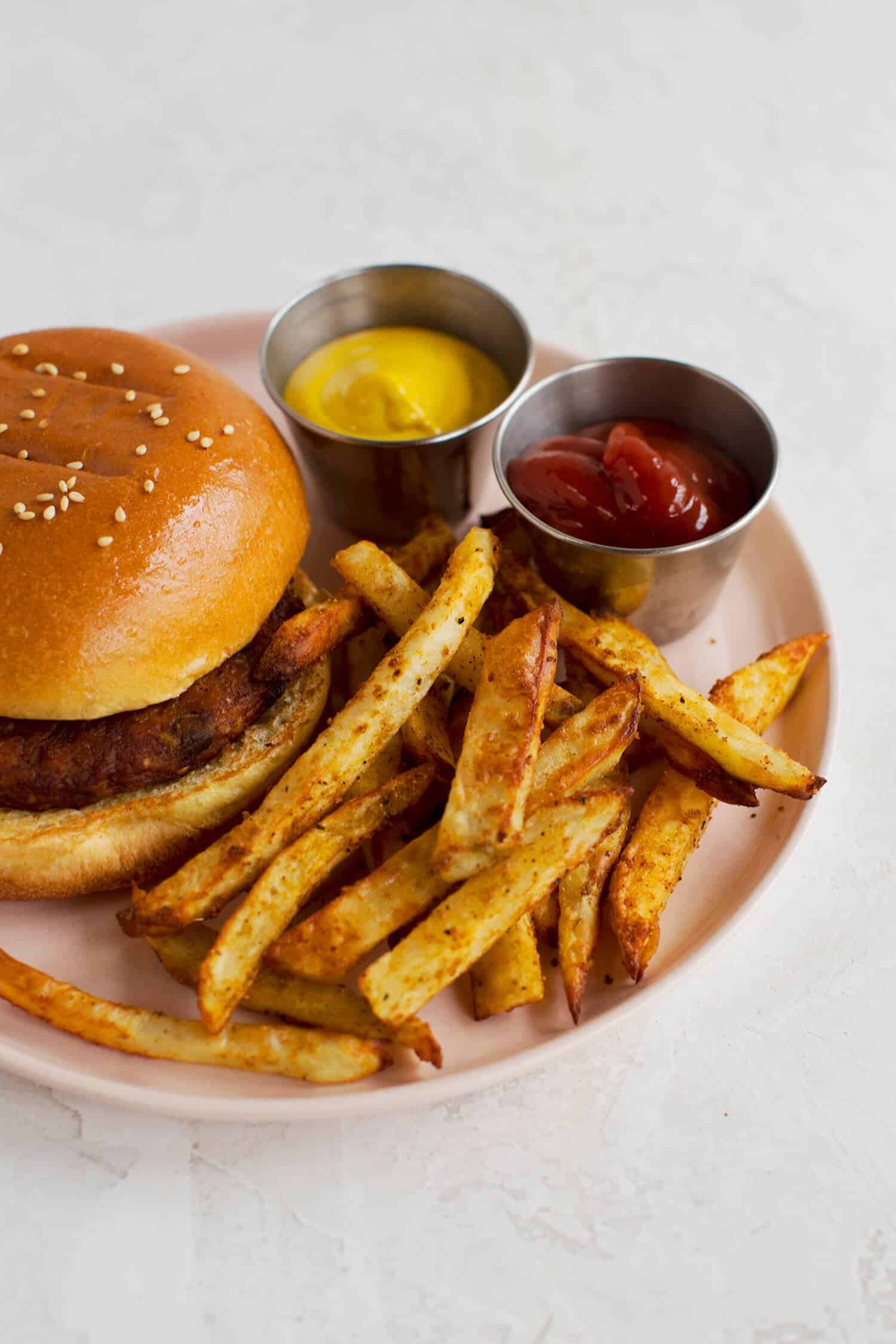 a pink plate with a hamburger, air fried seasoned french fries, a cup of mustard, and a cup of ketchup