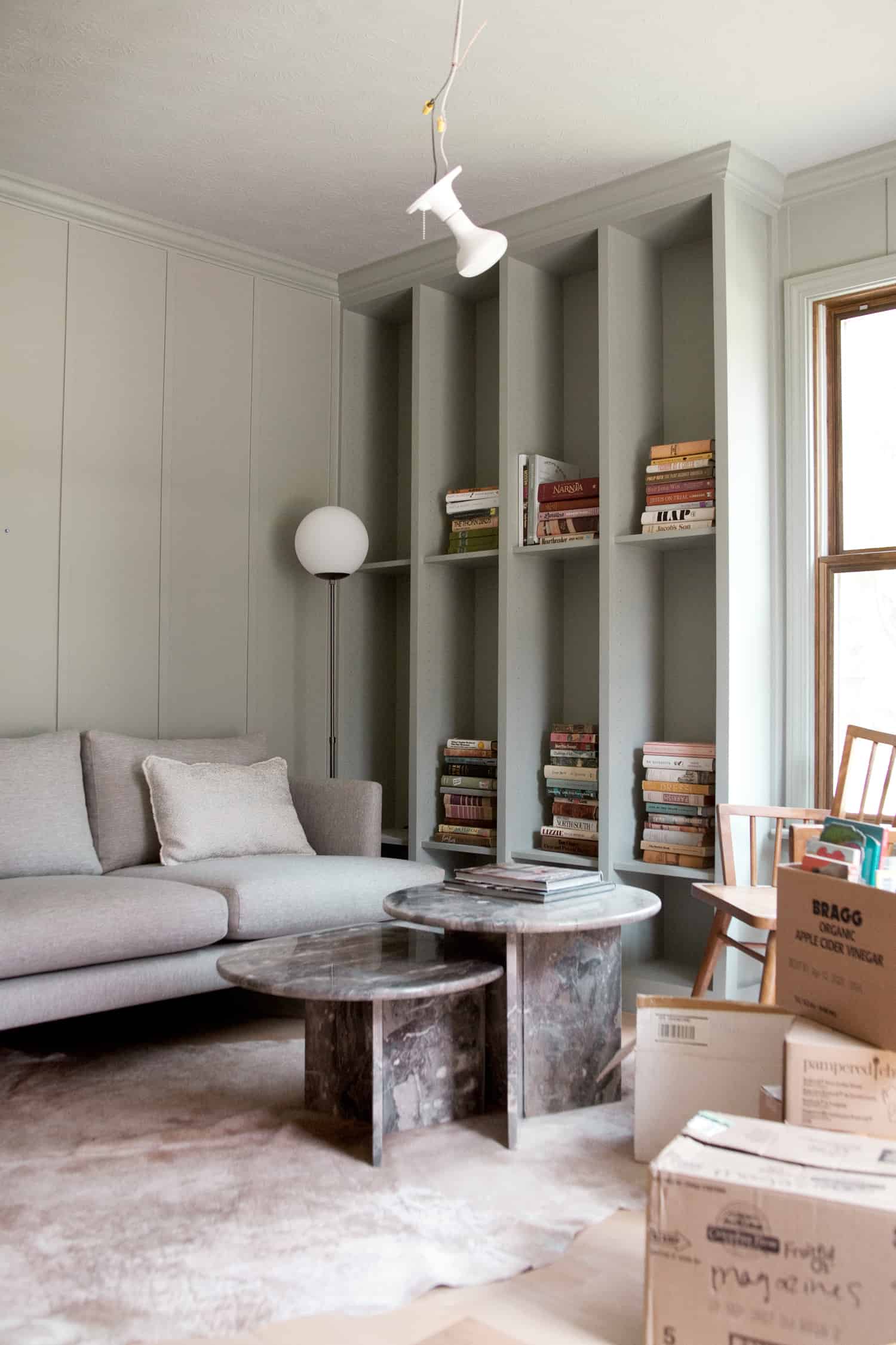 finished bookcase with piles of books on it, light fixture hanging from ceiling, boxes all over floor, gray coach, and coffee table