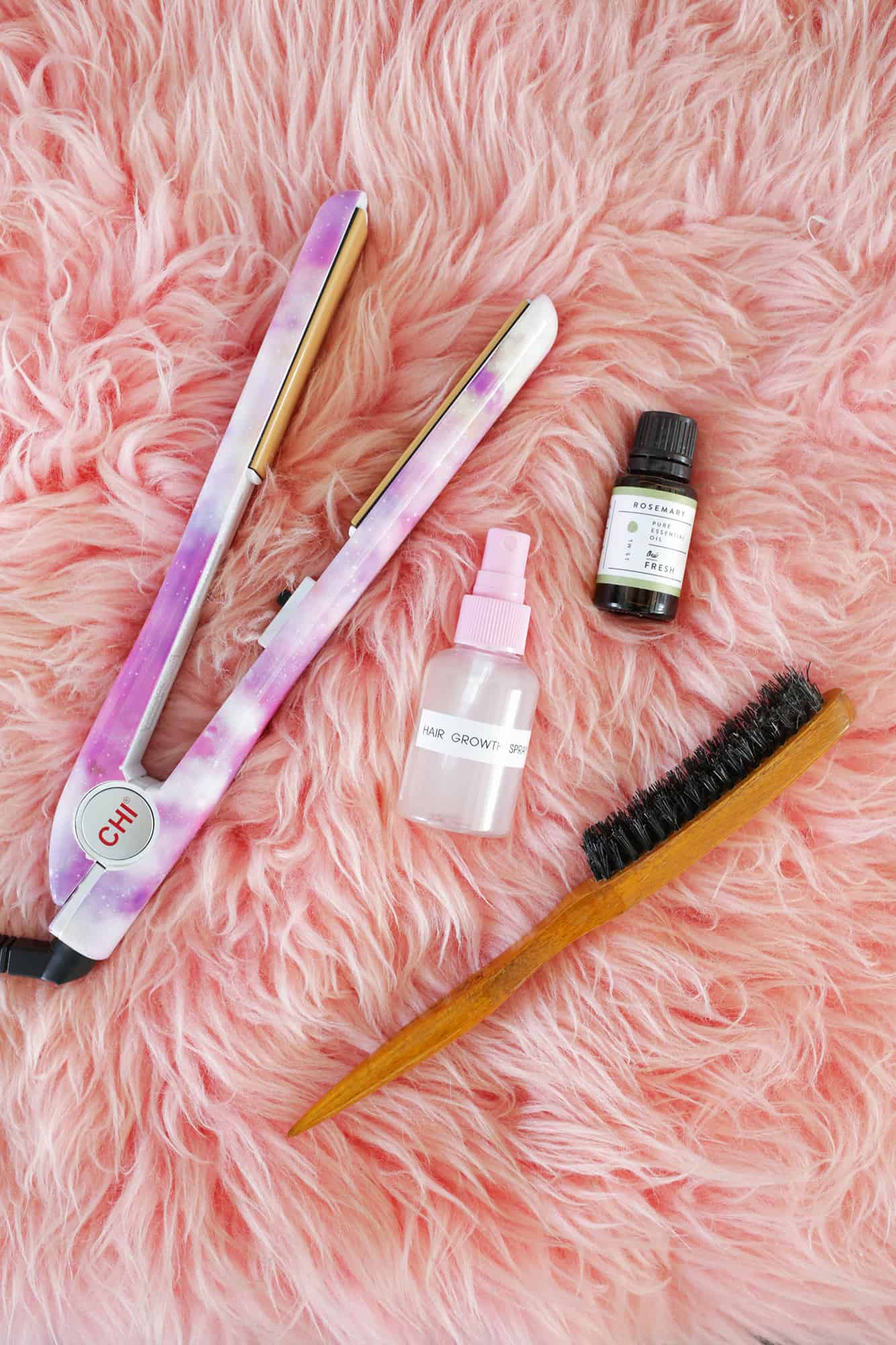 a curling iron, hair growth spray, Rosemary essential oil, and a hairbrush on a pink fluffy rug