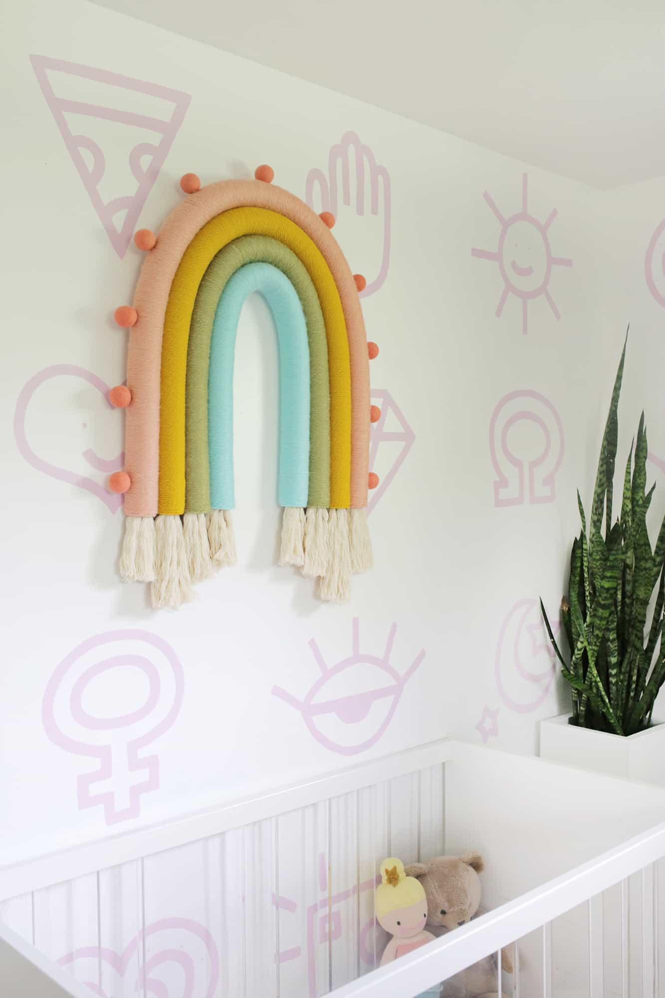 nursery with a white crib, plant, and a rainbow wall hanging on the wall above the crib