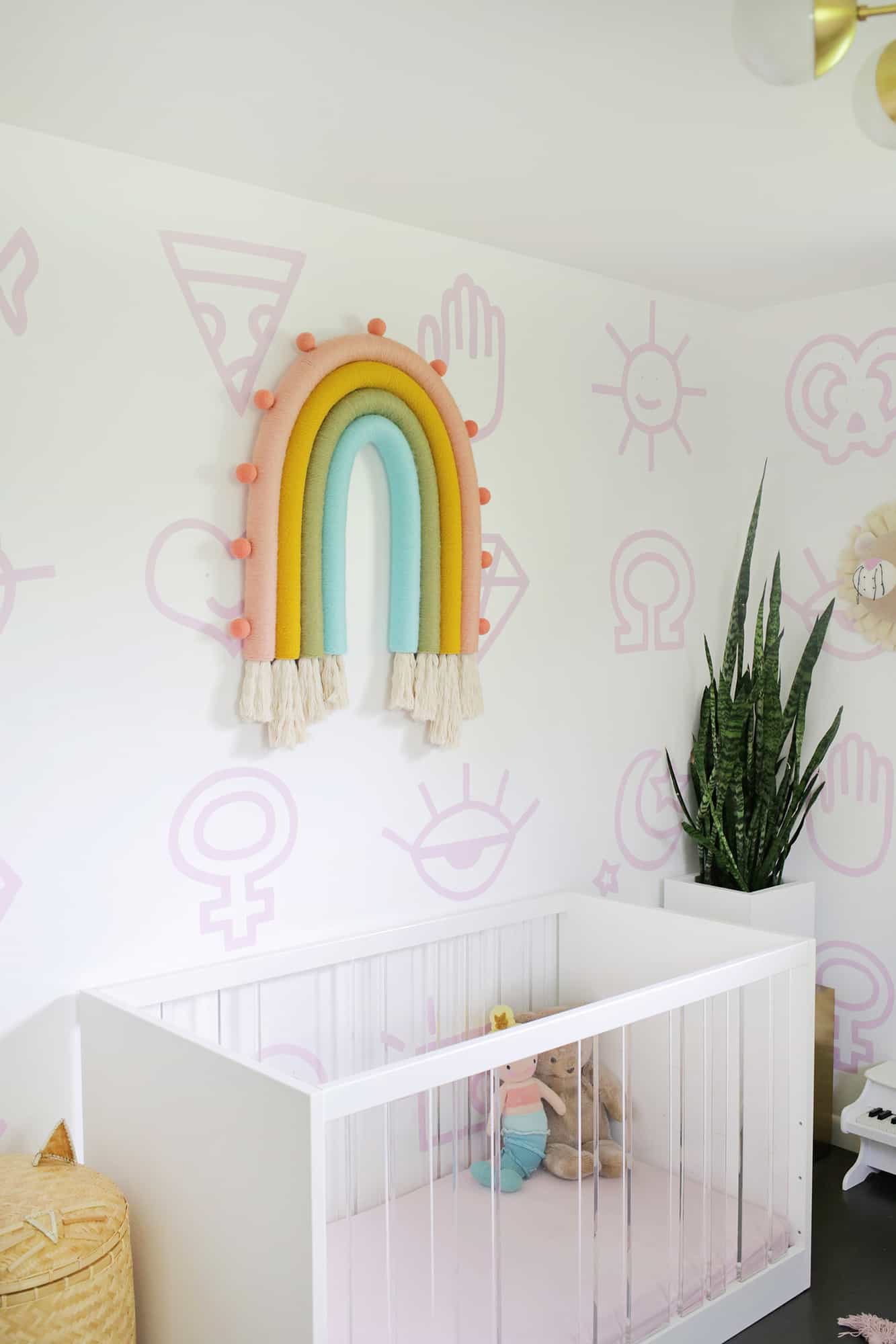  a nursery with a white crib, a plant, and a rainbow wall hanging above the crib