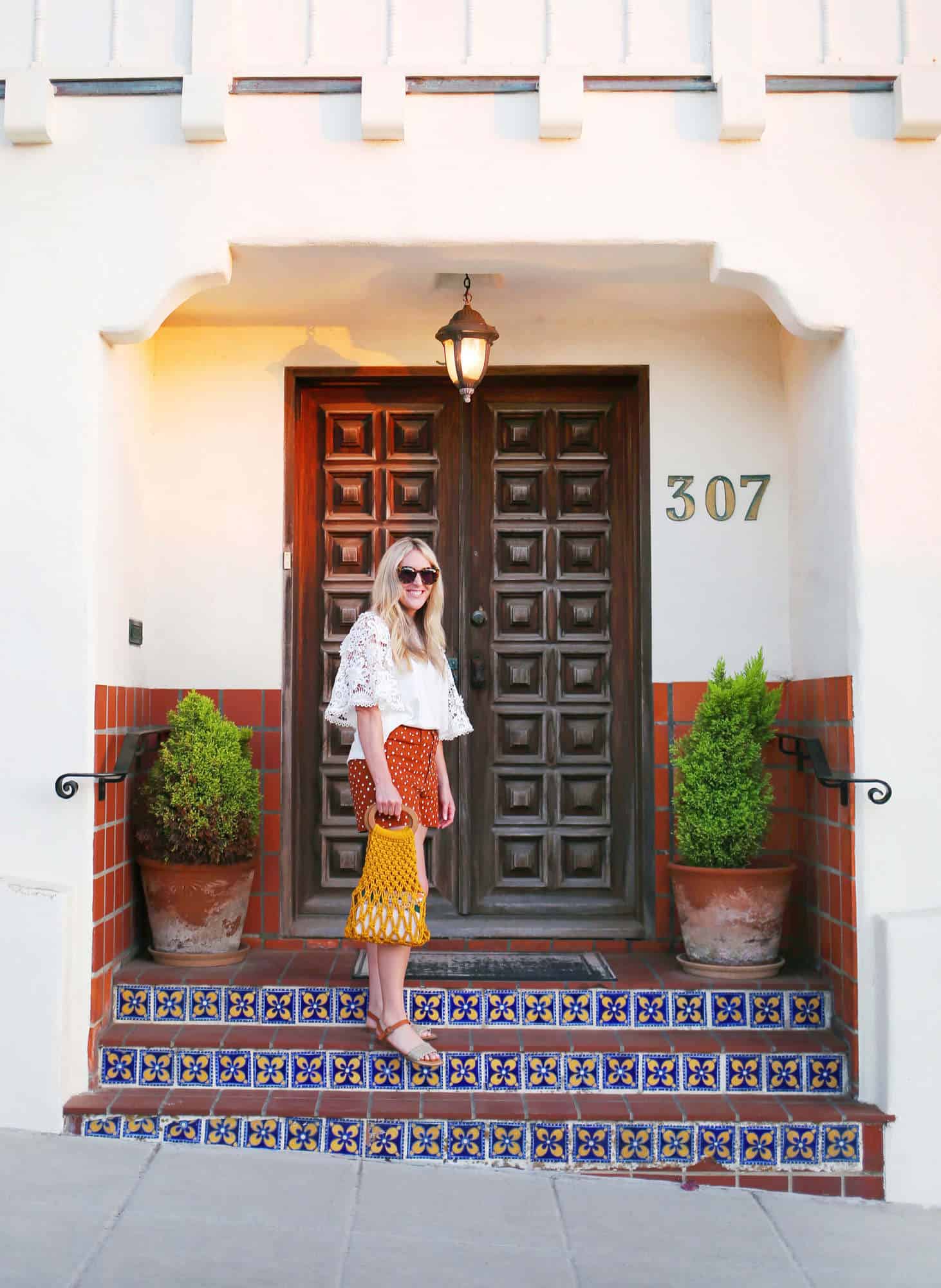 a blonde woman wearing sunglasses, a white long sleeve shirt, red shorts with white polka dots, and scandals standing on stairs in front of a house