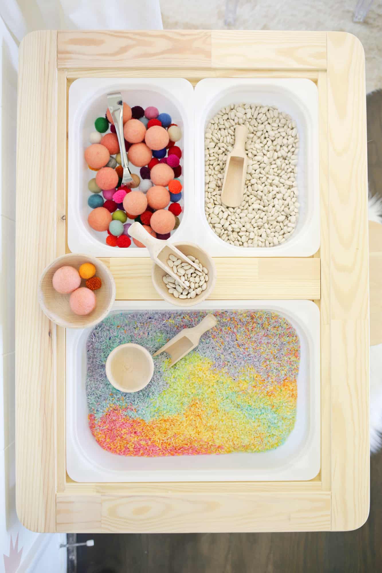 a white bin of colorful rice, a wooden scoop, and a wooden bowl, another white bin of colorful foam balls and tongs, and another white bin of beans and a wooden scoop in sensory table