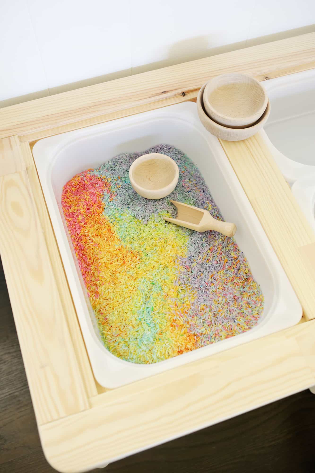 colorful rice, a wooden scoop and a wooden bowl in a white bin of the sensory table