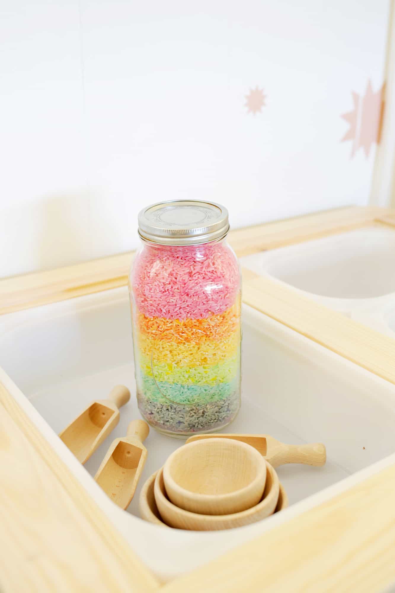 glass jar of colorful rice, 3 wooden scoops, and 3 wooden bowls in a white bin in sensory table