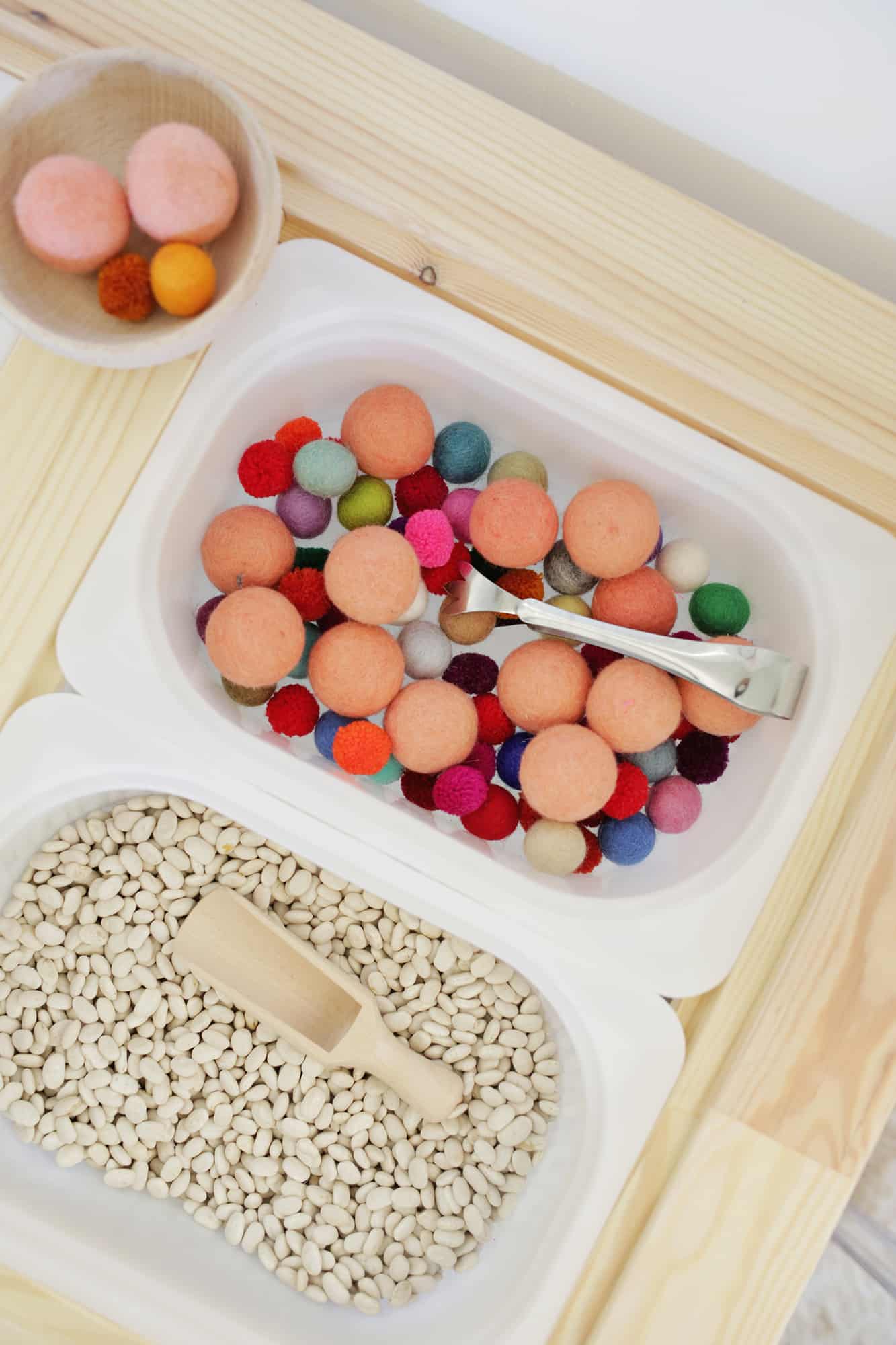 colorful foam balls with tongs in a white bin and beans and a wooden scoop in a white bin on sensory table