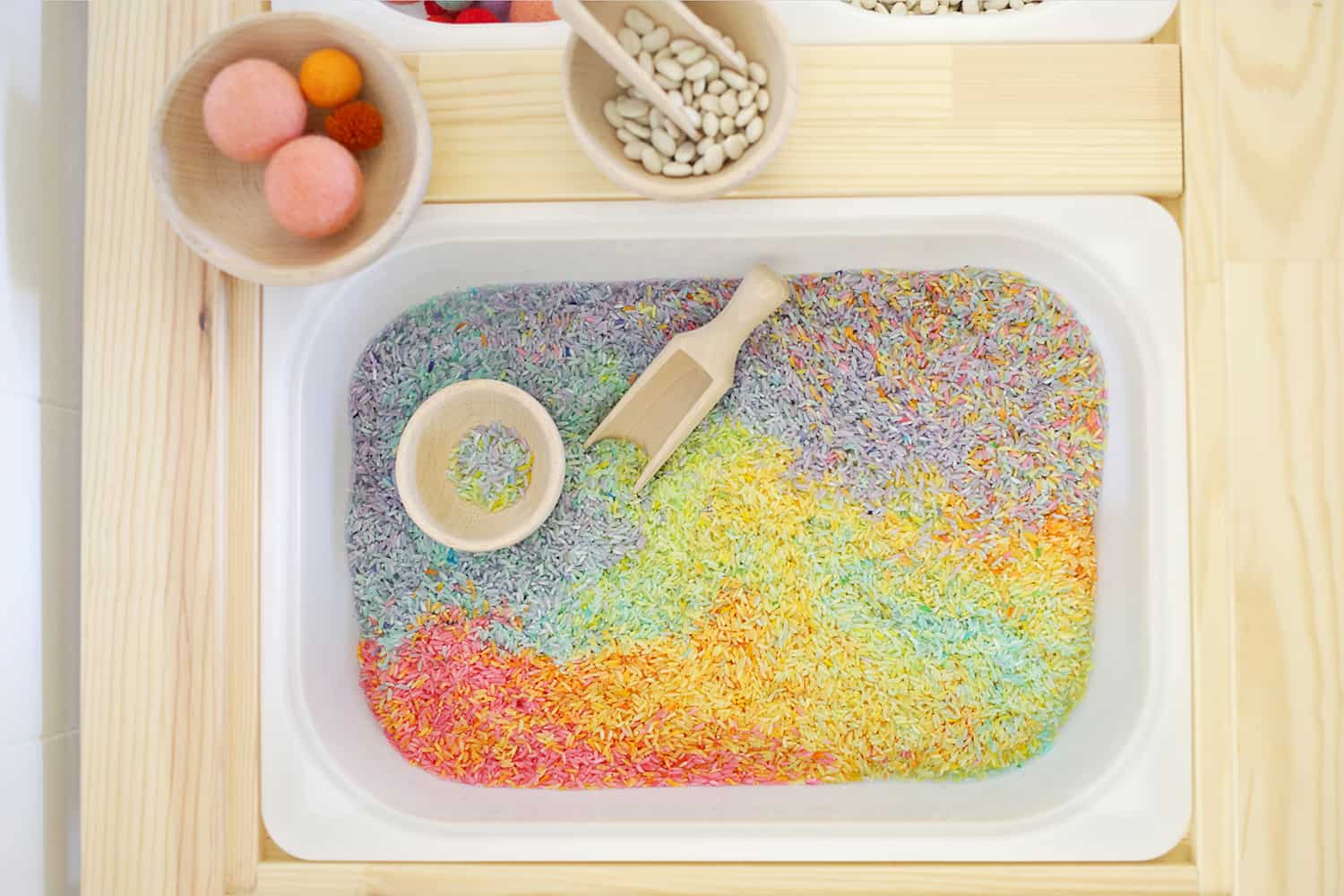 colorful rice, a wooden scoop, and a wooden bowl in a white bin in sensory table