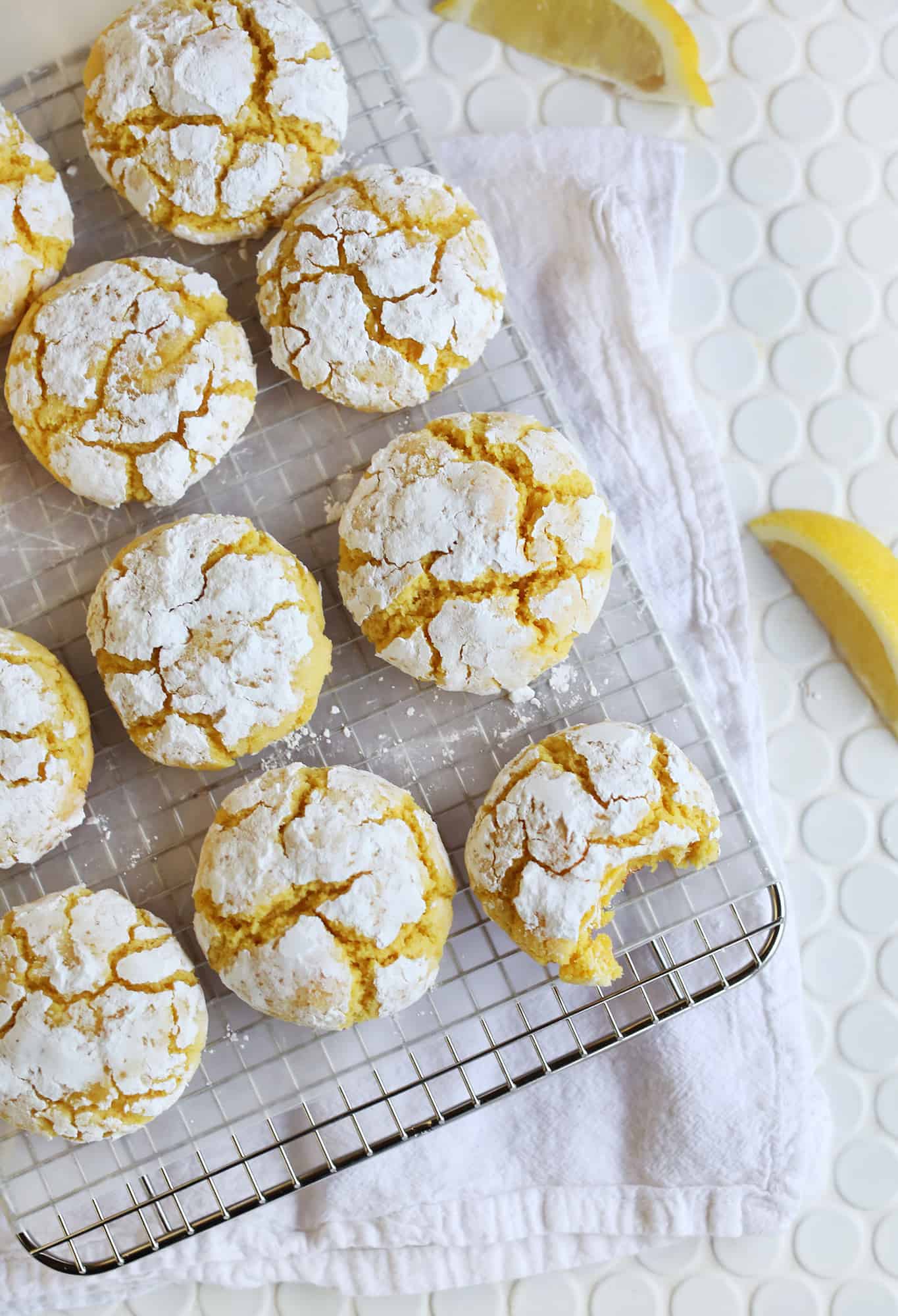 lemon burst crinkle cookies on a cooling rack with one cookie having bite taken out of it and lemons laying next to cooling rack