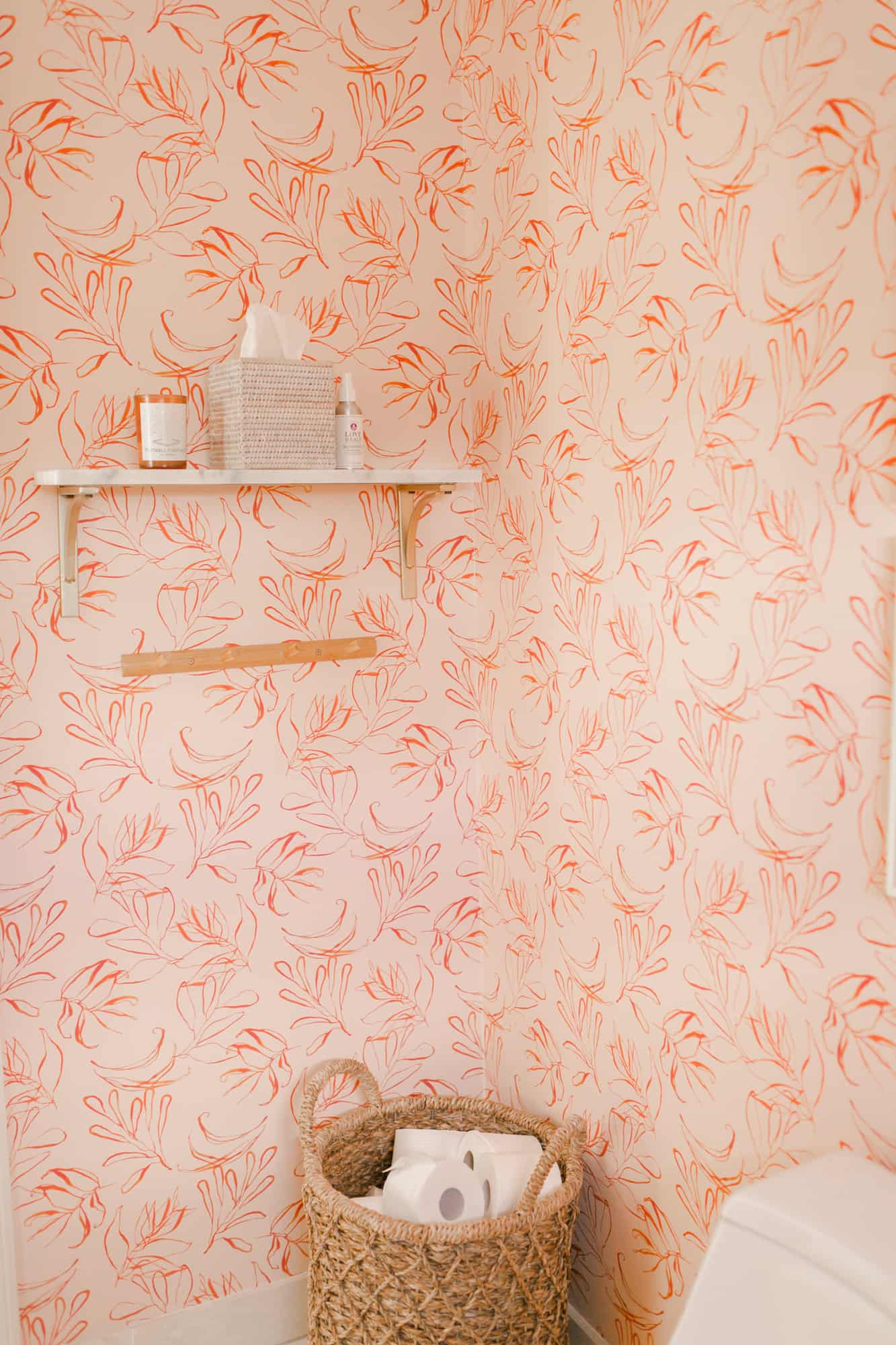 pink with flower wallpaper in a bathroom with basket of toilet paper on the floor and a white shelf with a candle and tissues on it