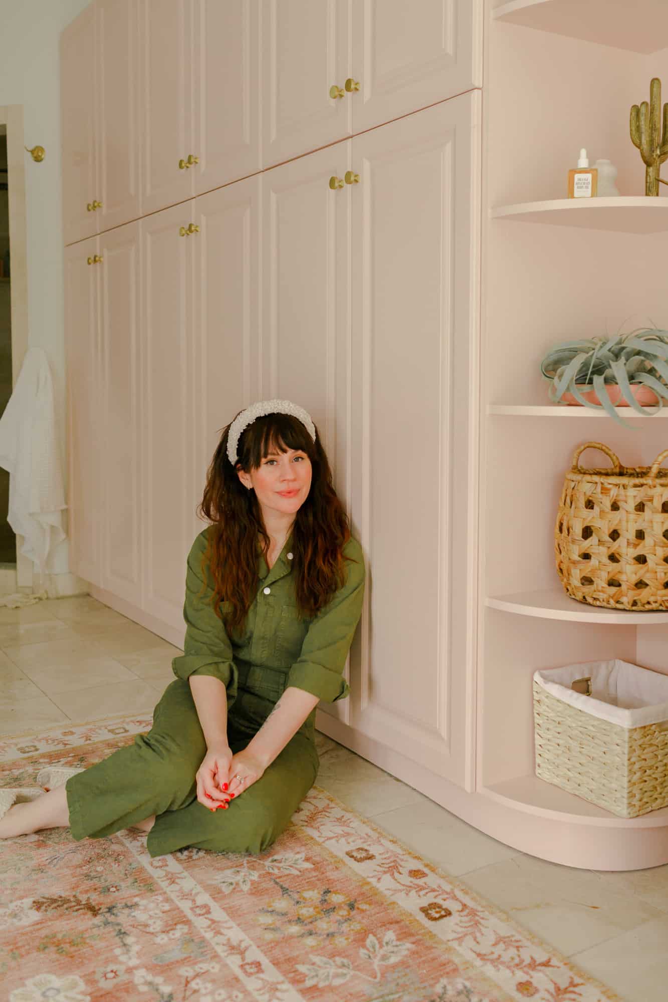 Elsie wearing  green jumpsuit with a white headband sitting on the floor in front of pink cabinets