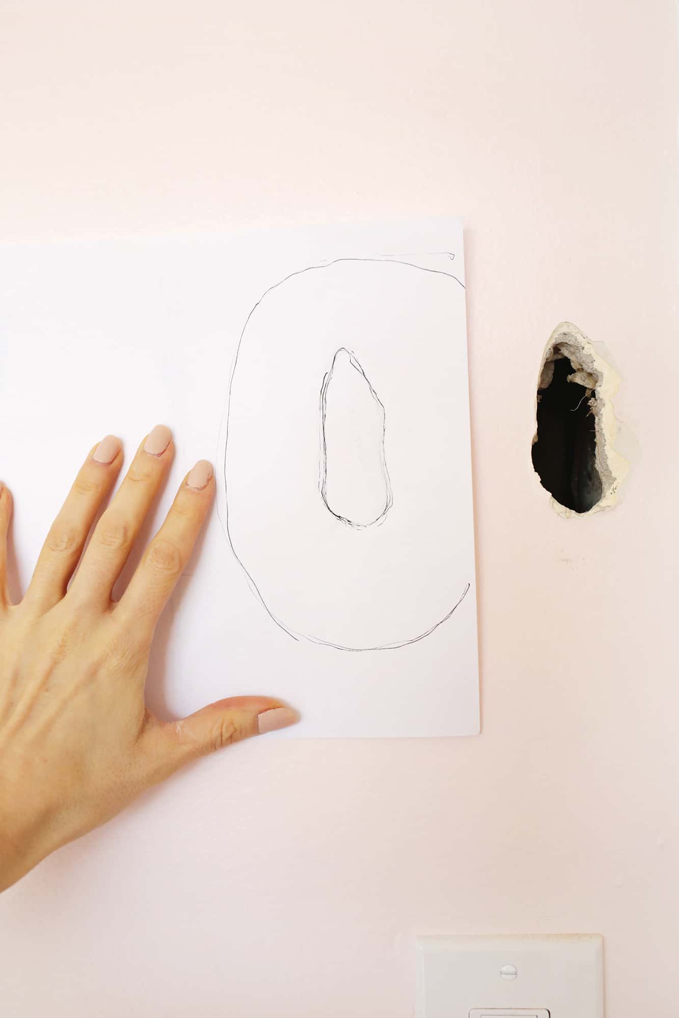 someone holding an outline of the hole on a white piece of paper next to the hole in the wall