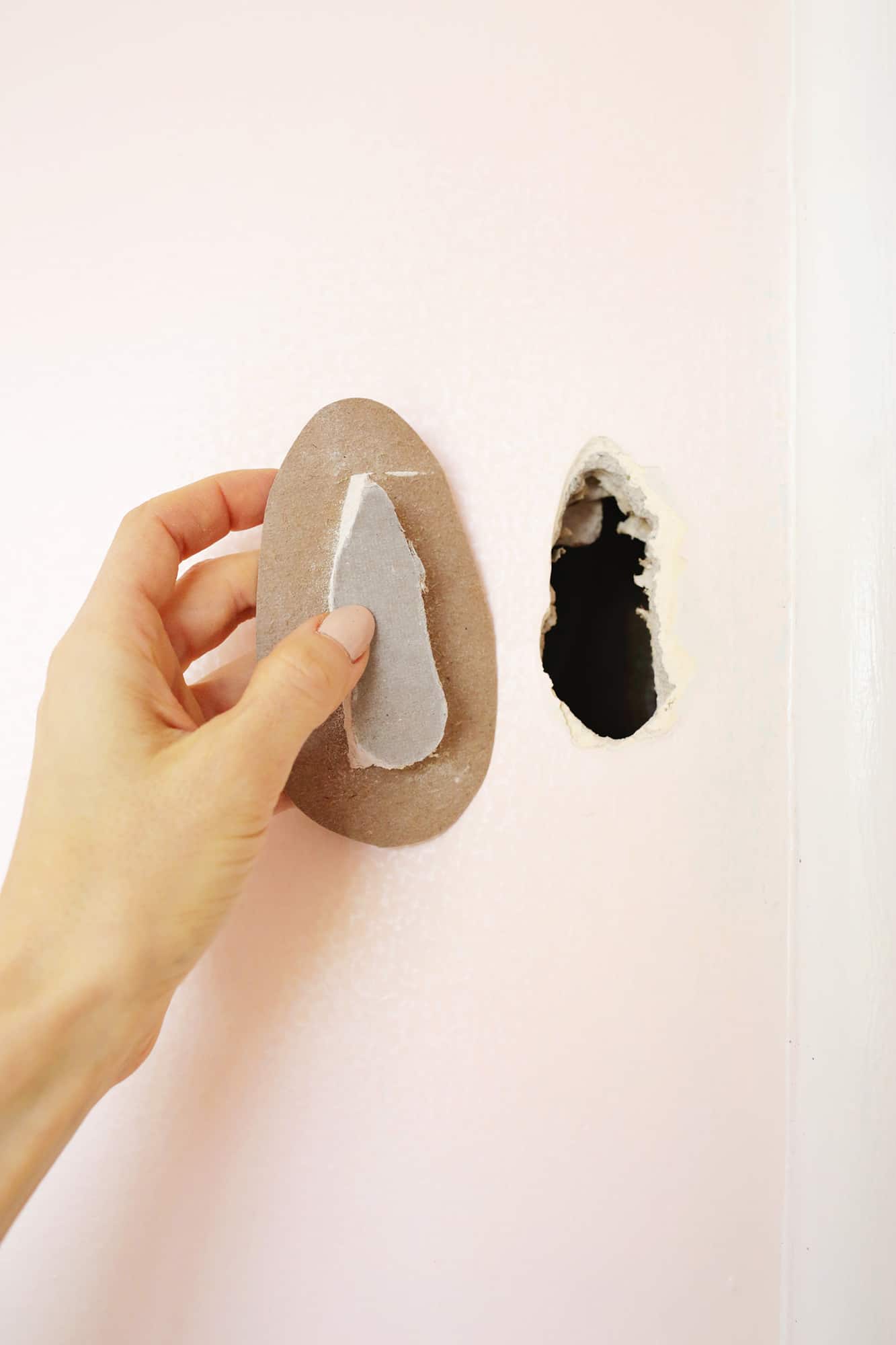 a pink wall with a hole in it and someone holding the part of the drywall that came out