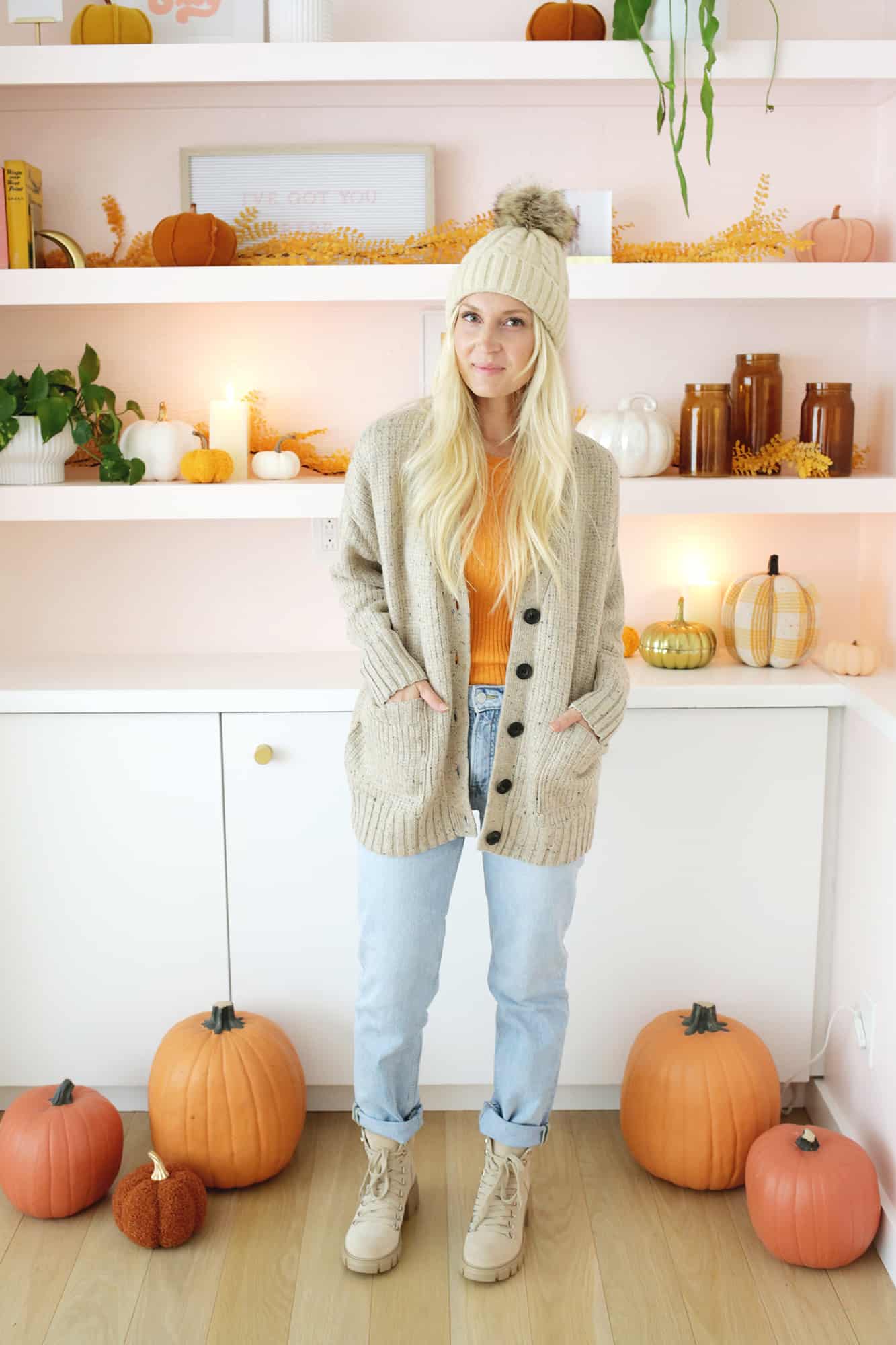 women wearing an oversized cardigan, hat with pom pom, jeans, and boots in front of fall styled bookshelf
