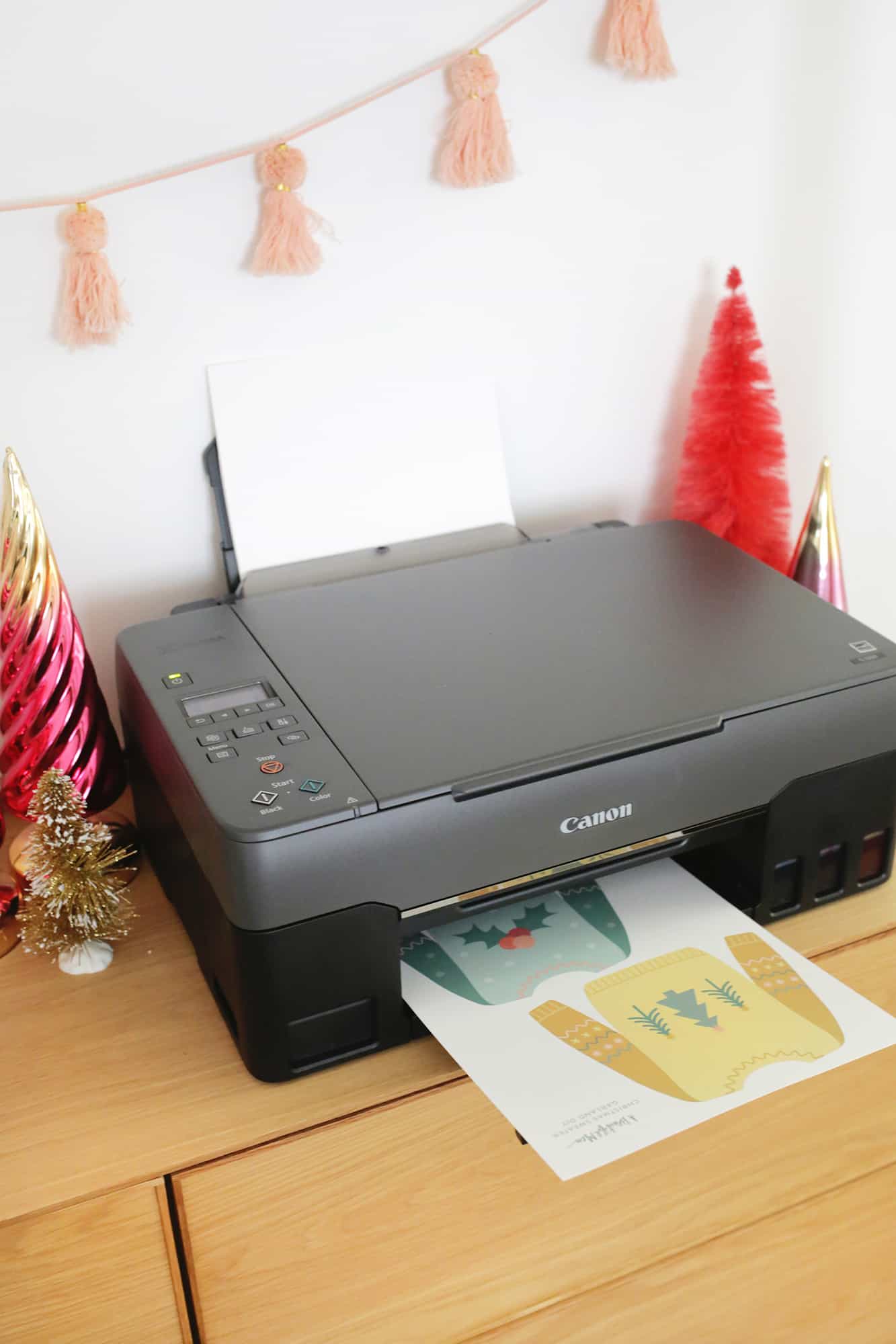 printer printing images of chistmas sweaters on paper with Christmas decor around the printer