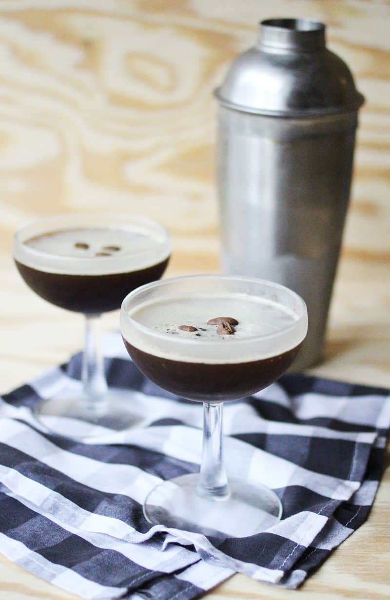 Two espresso martinis on a blue checkered towel in front of a cocktail shaker. 