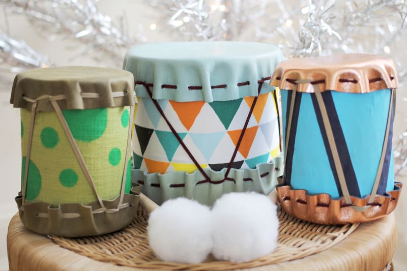 DIY Drums one is beige with a light green center with dark green polka dots, the second is bigger and light blue with brown lacing with white,  blue, turquoise, and orange, and black upside down triangle center, and the last copper with brown and beige threading with a blue and light blue stripe center, with drum sticks with two big white pom poms at the end.