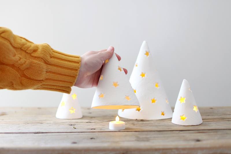 4 porcelain holiday tree lights with someone lifting one up to reveal a tea light under it