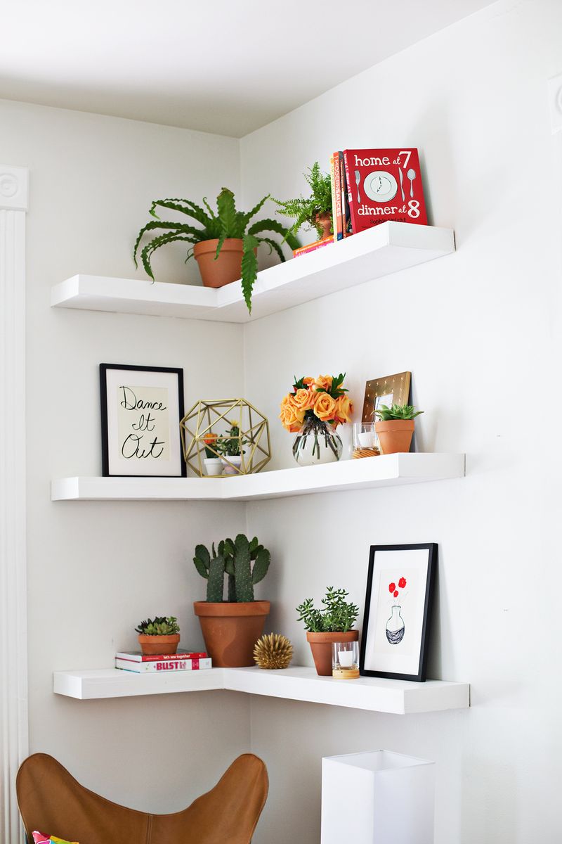 3 white floating corner shelves with books, plants, and decor on them