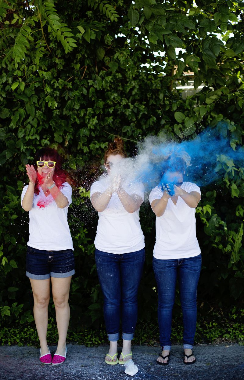 3 women in white t-shirts and jeans clapping colored powder between their hands