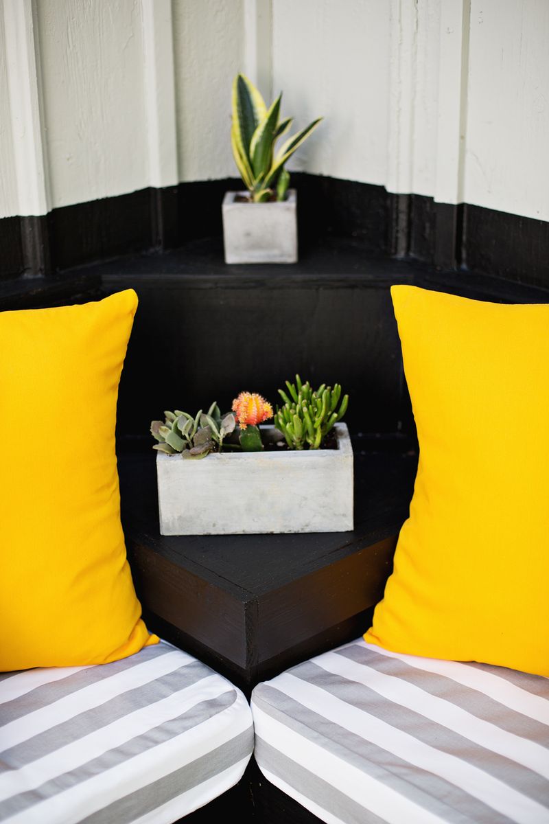 corner of the black bench with a gray and white seat cushion and yellow pillows. 2 plants in corner of bench