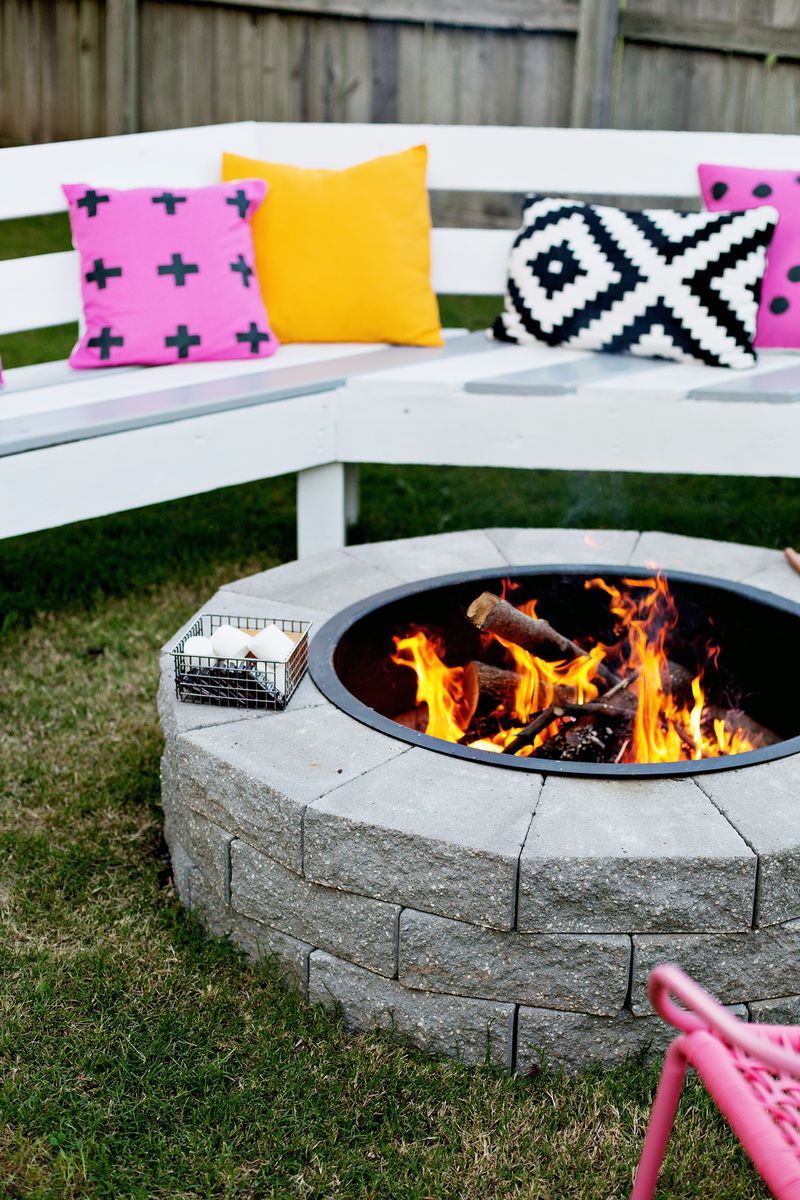 firepit with fire burning and a basket of marshmallows on it with bench an pillows surrounding firepit