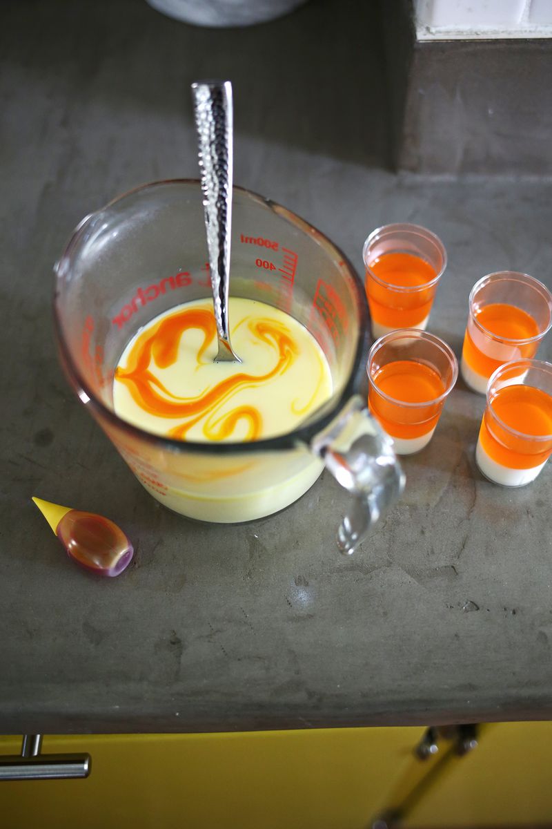 a mixing cup with milk mixture and swirls of orange food coloring in it with a spoon sticking out and 4 shot glassed with milk mixture and gelatin in it