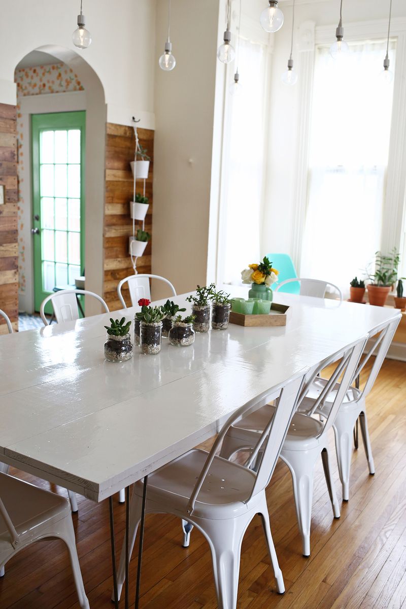 white painted dining room table with 8 chairs with 7 plants, a wood tray, a vase with flowers, and 4 green glasses on top