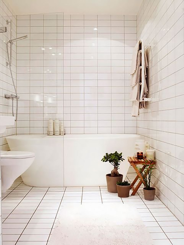 White Stack bond subway tile in a bathroom with a white tub, white toilet, 3 potted plants, and a wooden table with candles on it