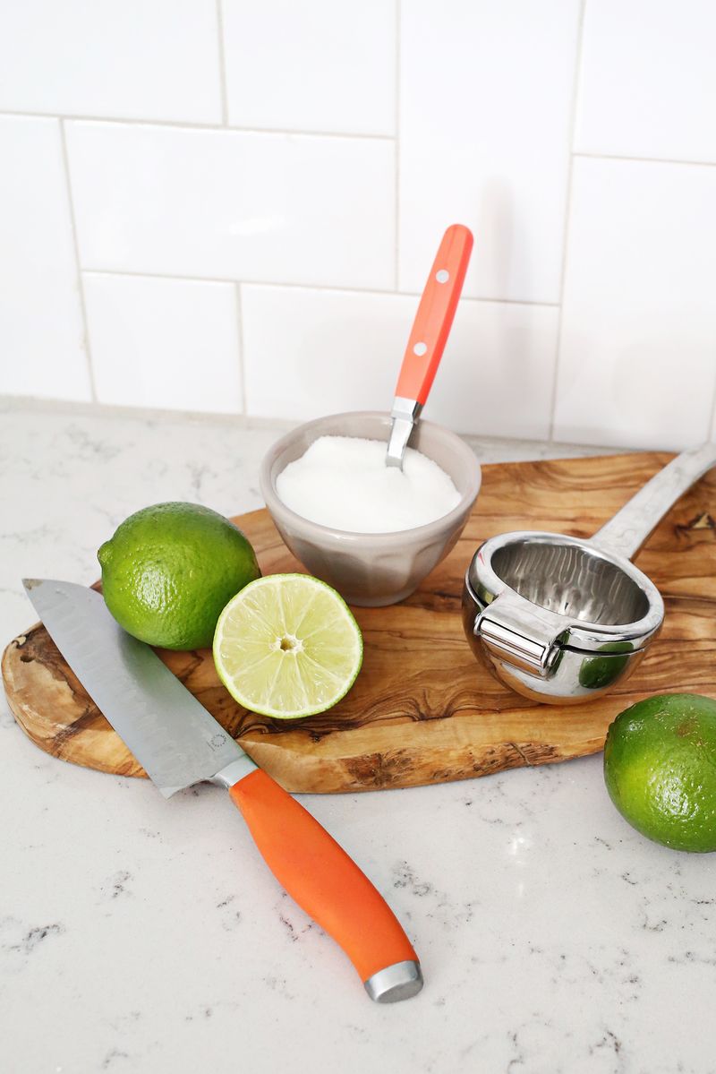 2 whole limes and 1 half lime on a cutting board with a cup of sugar and a lime press