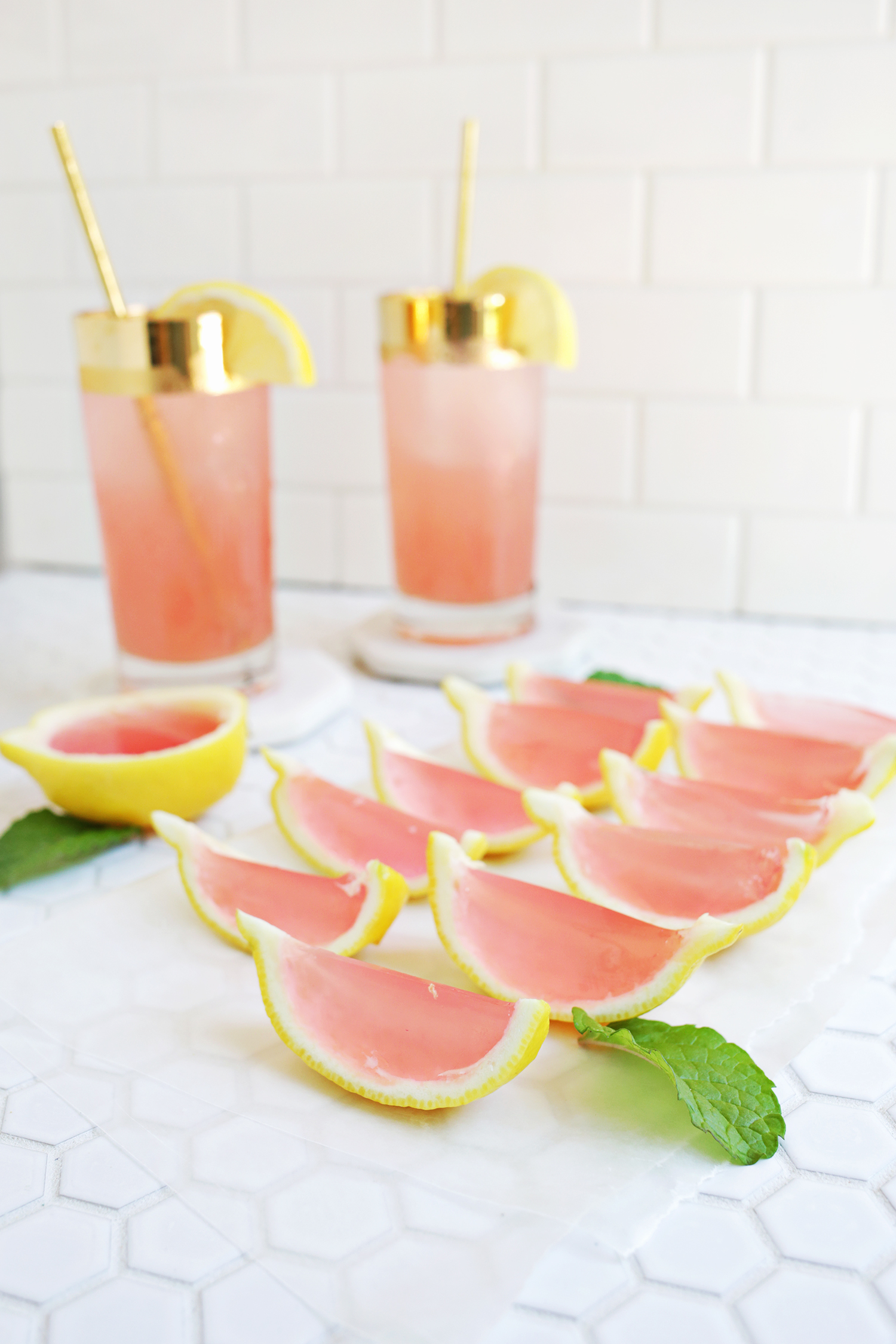 Pink Lemonade Jello Shots in 2 tall glasses with yellow straws with pink lemon halves in front of them