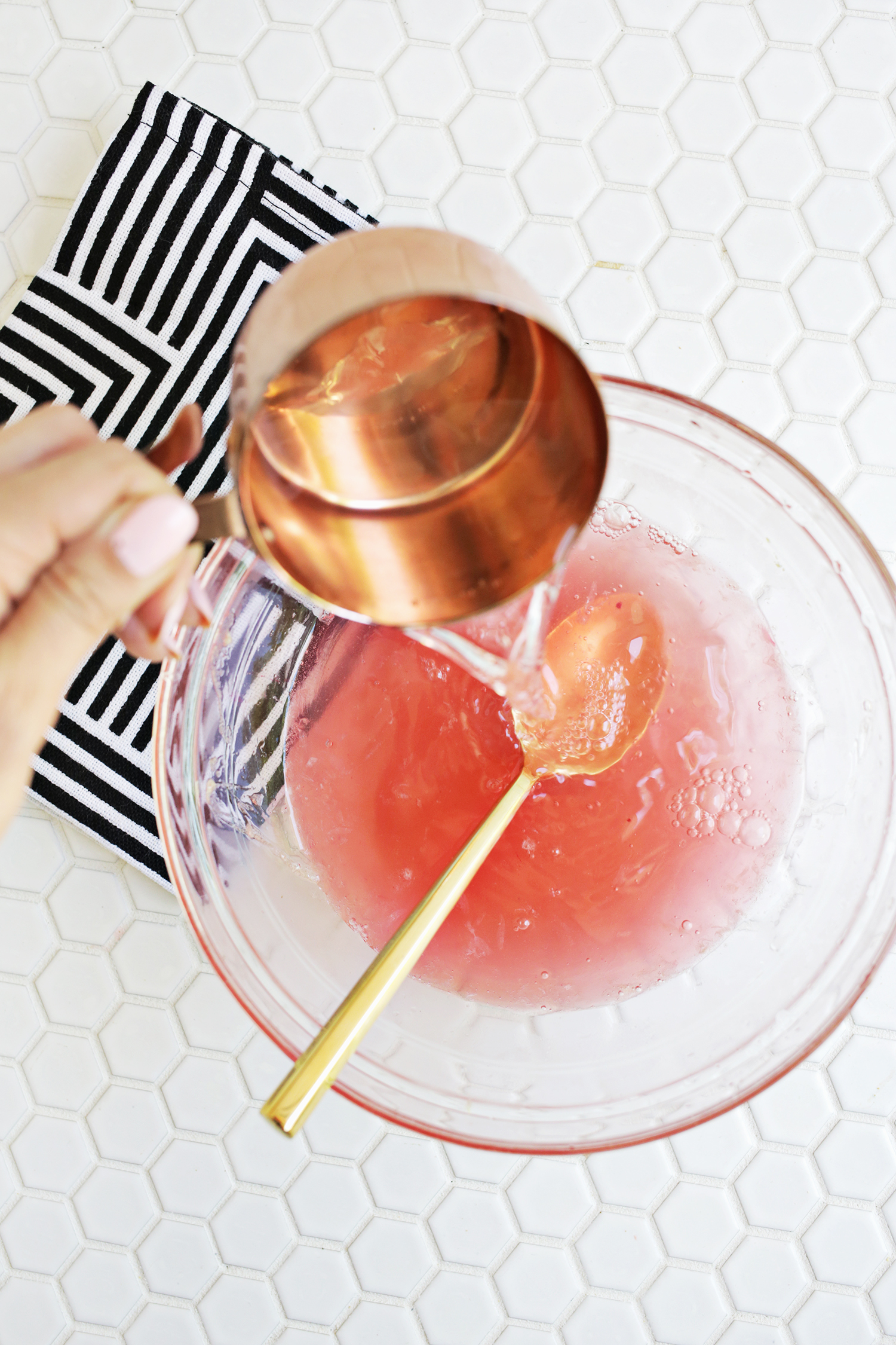 someone pouring pink lemonade from a cooper mixing cup into a glass mixing bowl with a spoon in it