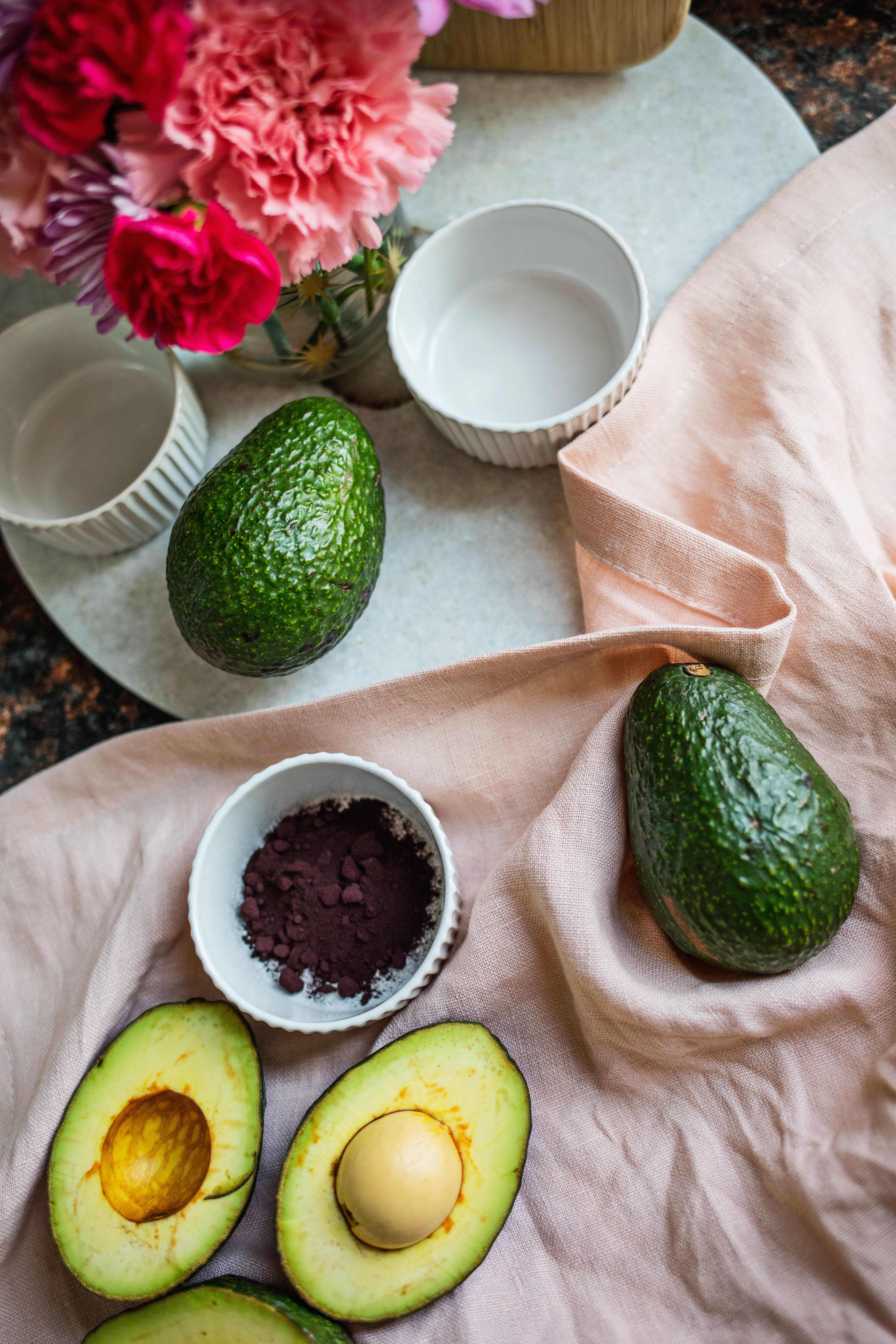 pink fabric with a whole avocado, a cut in half avocado, and a bowl of purple powder on it