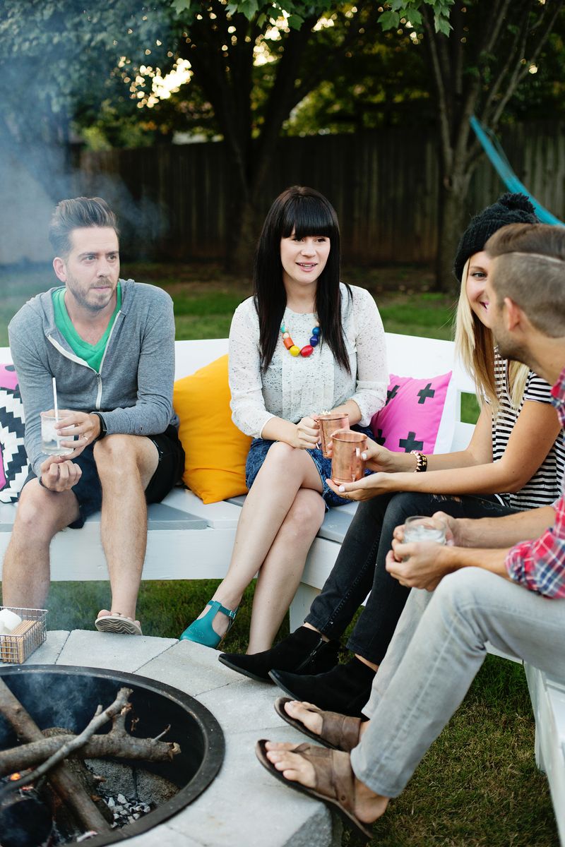 2 couples talking and sitting on bench around fire pit holding cups