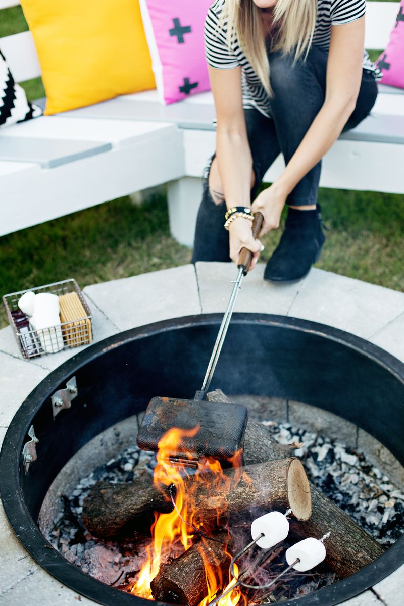 a woman holding a screwer over the fire in the fire pit with a basket of more supplies next to her