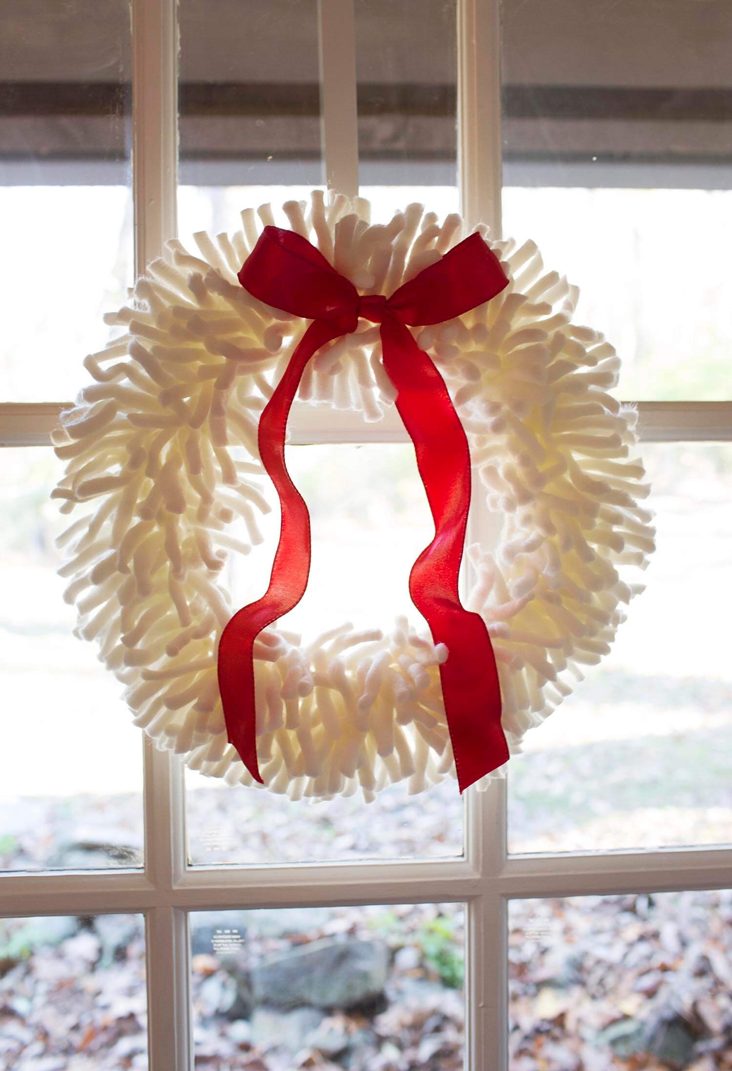 white yarn wreath with a red ribbon hanging on a window 