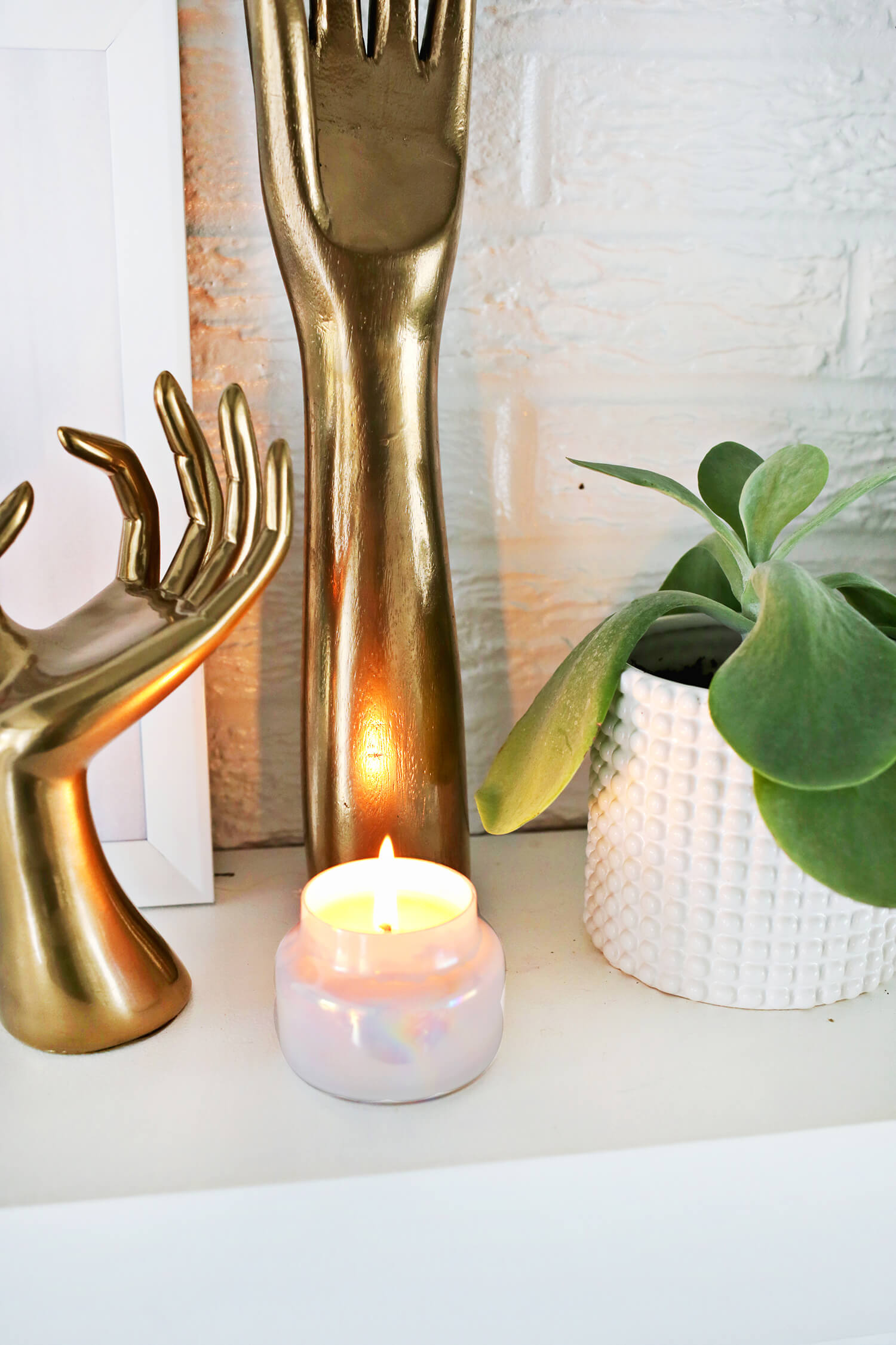 a beeswax candle lite by a plant and 2 arm and hand statues